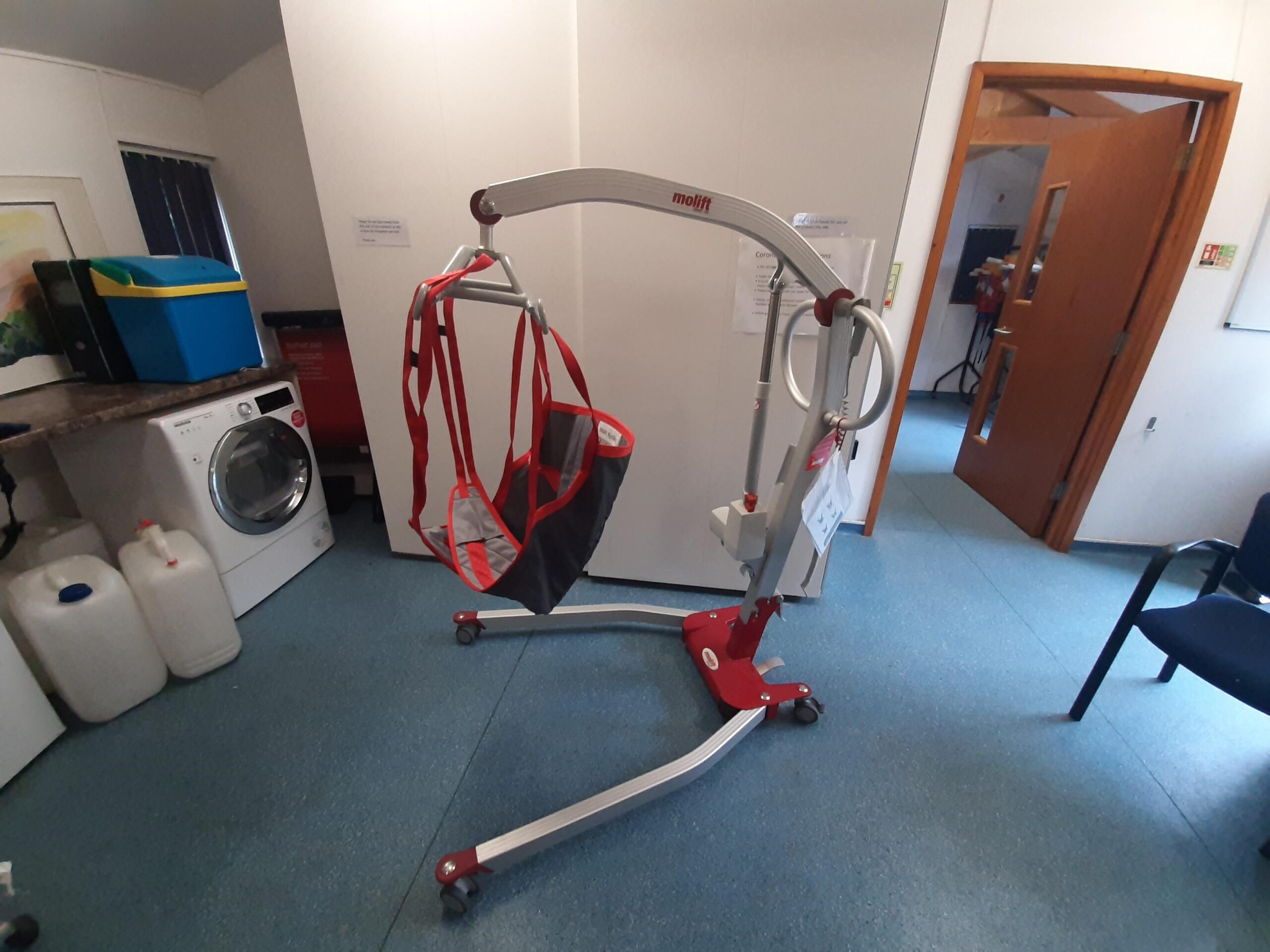 Molift Hoist with sling in position ready to use. In room with blue flooring and white walls with a wooden door.