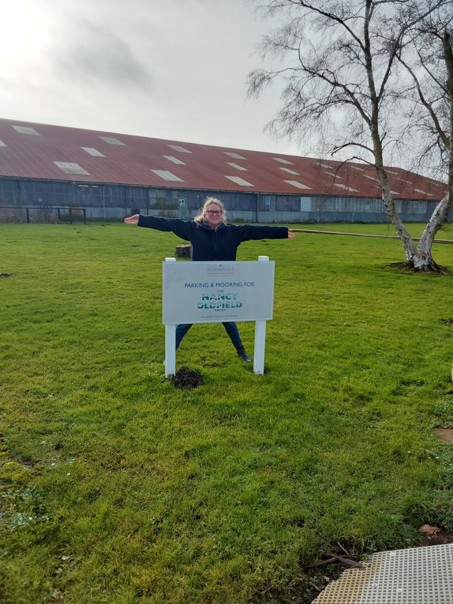A lady standing in star shaped stance behind a white sign on two legs, on the grass. The sign reads Nancy Oldfield Trust parking and mooring.